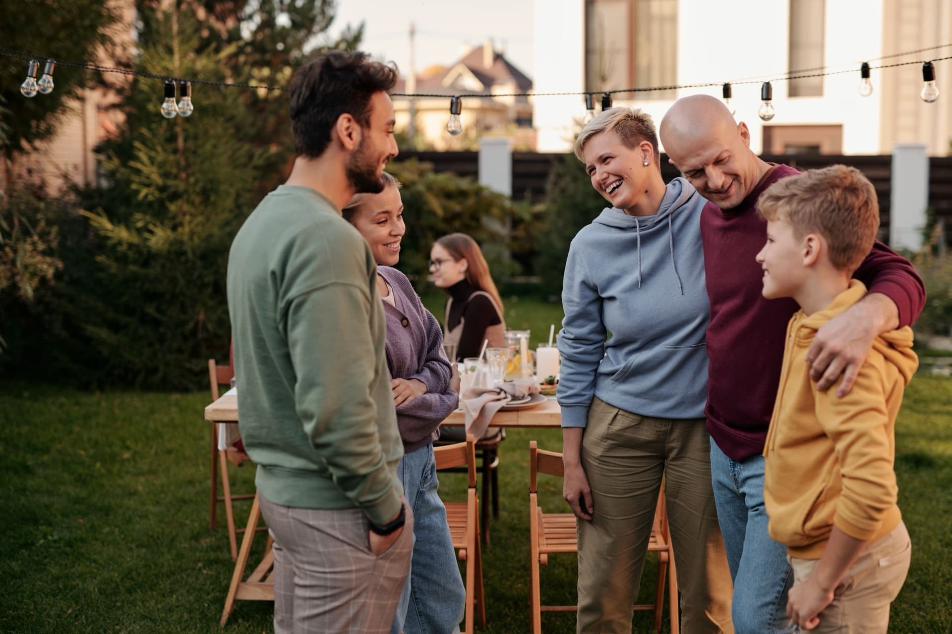 Family and friends laughing together at an outdoor table in a backyard strung with festoon lights