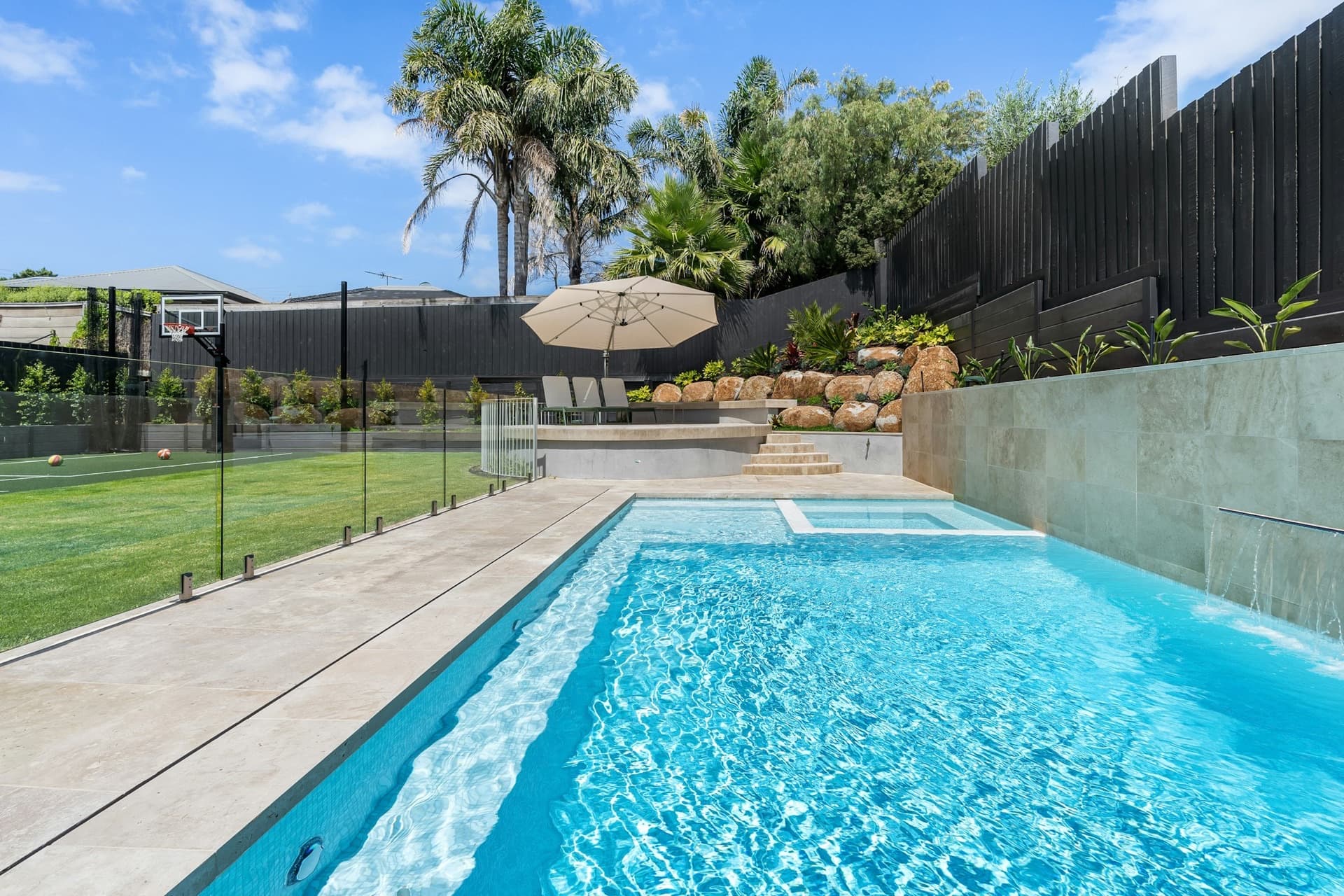 Backyard swimming pool behind a frameless glass safety fence with a green lawn and basketball hoop beyond