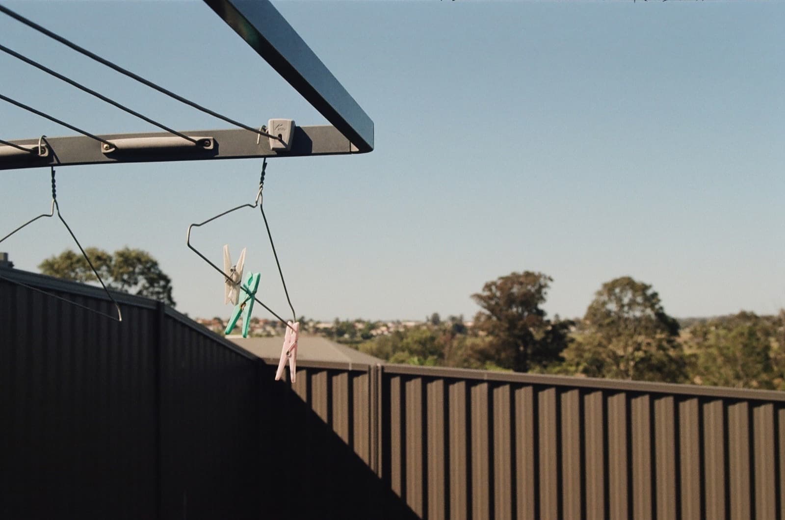 Rotary clothesline arm with pegs and hangers over a Colorbond fence, gum trees in the distance